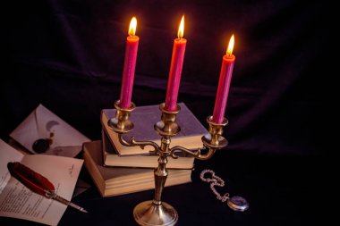 Bronz şamdanlı şarap rengi mumlar, antika kitaplar, tüy kalem ve koyu mavi arka planda eski zarflar.