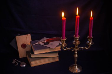 Bronz şamdanlı şarap rengi mumlar, antika kitaplar, tüy kalem ve koyu mavi arka planda eski zarflar.