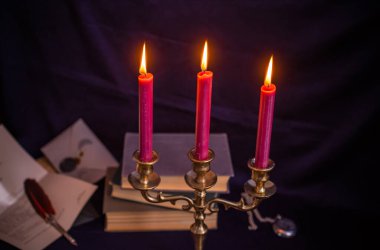 Bronz şamdanlı şarap rengi mumlar, antika kitaplar, tüy kalem ve koyu mavi arka planda eski zarflar.