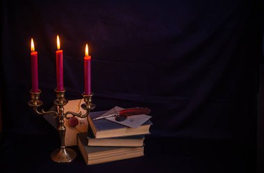 Bronz şamdanlı şarap rengi mumlar, antika kitaplar, tüy kalem ve koyu mavi arka planda eski zarflar.