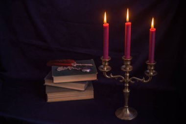 Mistik bronz bir üçlü mum tutacağı, yanan mumlar, antik kitaplar ve yazı kalemi. Sıkıntılı karanlık doku dengesiz ve gotik bir atmosfer yaratıyor.