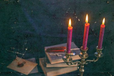 Mistik bronz bir üçlü mum tutacağı, yanan mumlar, antik kitaplar ve yazı kalemi. Sıkıntılı karanlık doku dengesiz ve gotik bir atmosfer yaratıyor.