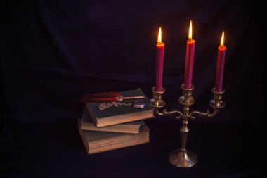 Mistik bronz bir üçlü mum tutacağı, yanan mumlar, antik kitaplar ve yazı kalemi. Sıkıntılı karanlık doku dengesiz ve gotik bir atmosfer yaratıyor.