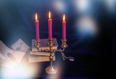 Mistik bronz bir üçlü mum tutacağı, yanan mumlar, antik kitaplar ve yazı kalemi. Sıkıntılı karanlık doku dengesiz ve gotik bir atmosfer yaratıyor.