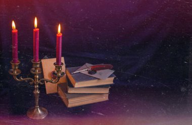 Mistik bronz bir üçlü mum tutacağı, yanan mumlar, antik kitaplar ve yazı kalemi. Sıkıntılı karanlık doku dengesiz ve gotik bir atmosfer yaratıyor.