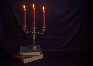 Mistik bronz bir üçlü mum tutacağı, yanan mumlar, antik kitaplar ve yazı kalemi. Sıkıntılı karanlık doku dengesiz ve gotik bir atmosfer yaratıyor.