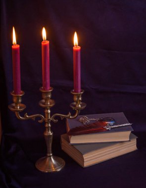 Mistik bronz bir üçlü mum tutacağı, yanan mumlar, antik kitaplar ve yazı kalemi. Sıkıntılı karanlık doku dengesiz ve gotik bir atmosfer yaratıyor.