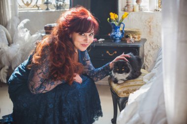 50 yaşında güzel bir kadın, kızıl saçlı, zarif mavi dantelli bir elbise giyiyor, lüks klasik bir koltukta oturuyor ve tüylü bir kedi tutuyor.