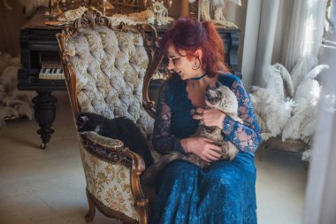 50 yaşında güzel bir kadın, kızıl saçlı, zarif mavi dantelli bir elbise giyiyor, lüks klasik bir koltukta oturuyor ve tüylü bir kedi tutuyor.