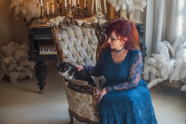 50 yaşında güzel bir kadın, kızıl saçlı, zarif mavi dantelli bir elbise giyiyor, lüks klasik bir koltukta oturuyor ve tüylü bir kedi tutuyor.