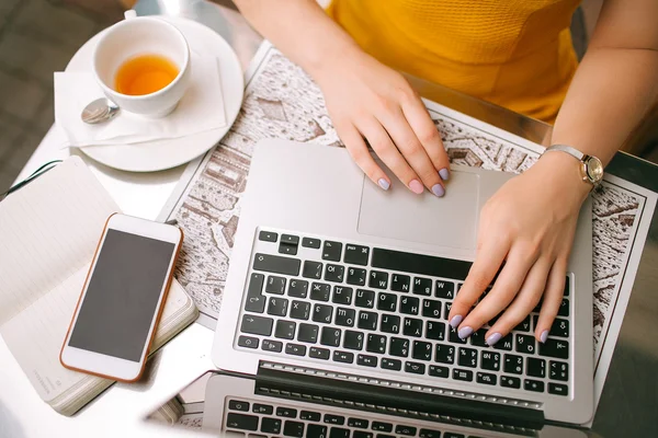 Manos sobre el portátil con el bloc de notas del teléfono inteligente y té en la cafetería — Foto de Stock