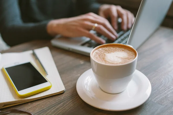 Manos del hombre en el ordenador portátil con café y smartphone con bloc de notas — Foto de Stock