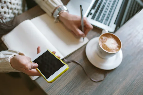 Руки женщины с ручкой и блокнотом, держащей смартфон с кофеином — стоковое фото