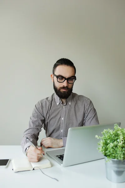 Homem com barba em óculos tomando notas com laptop e bloco de notas — Fotografia de Stock