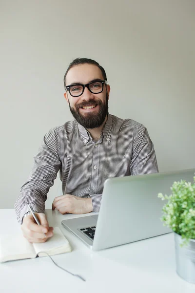 Homem sorridente com barba em óculos tomando notas com laptop notep — Fotografia de Stock