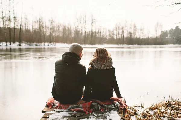 Young couple sitting on pier at frozen lake looking at each othe — Stock Photo, Image