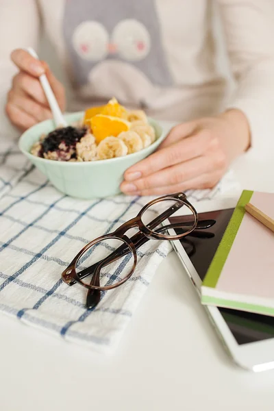 Kobieta jedzenie owsianka i owoce, notatnik tabletki i okulary na — Zdjęcie stockowe