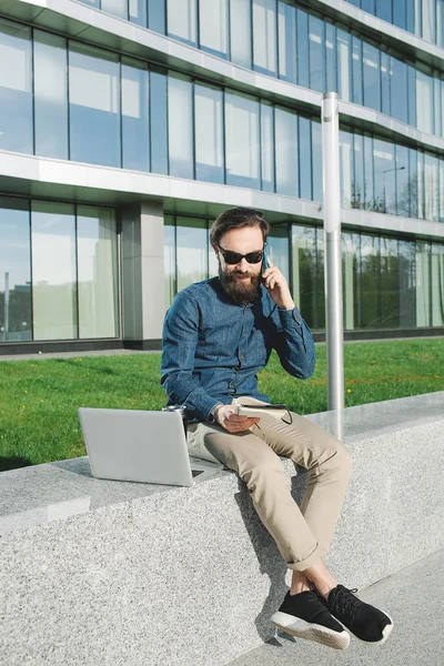 Joven hombre de negocios en gafas de sol con cafetera portátil hablando de teléfono —  Fotos de Stock