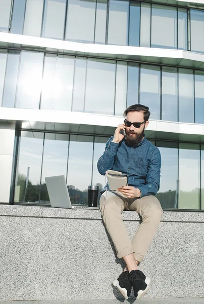 Joven hombre de negocios en gafas de sol con cafetera portátil hablando de teléfono —  Fotos de Stock