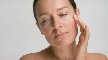 Pürüzsüz, sağlıklı bir cilde sahip genç bir kadının yakın plan güzel portresi, beyaz arka planda parmaklarıyla nazikçe yüzüne dokunur ve gülümser.