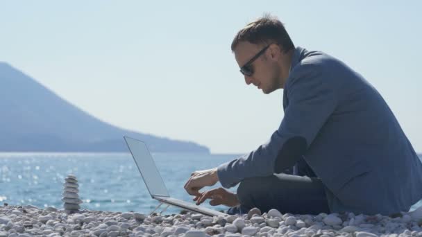 Empresário freelancer trabalhando remotamente no laptop na praia perto da pirâmide zen — Vídeo de Stock