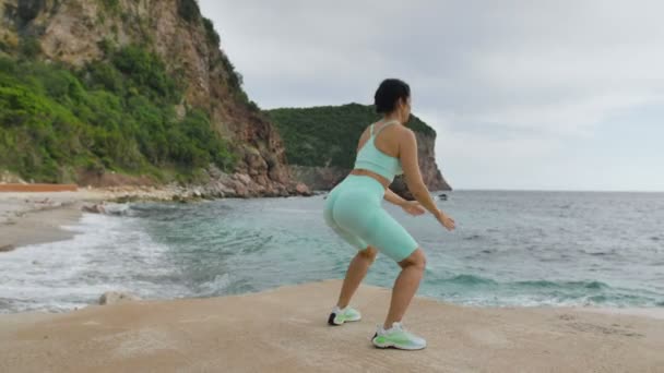 Ung attraktiv kvinna gör knäböj med hopp. Kvinnlig tränare som tränar utomhus på stranden. Sexig kvinna i sportkläder gör träning. — Stockvideo