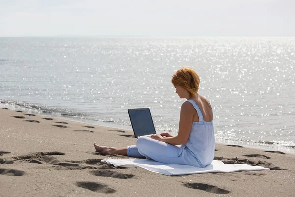 Redheaded girl working at a laptop sitting on the beach Stock Photo