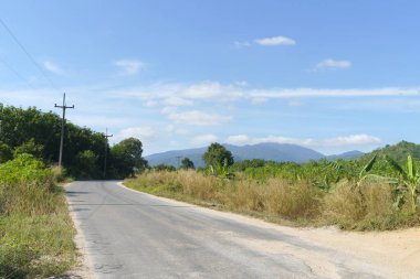 Dolambaçlı asfalt bir yol, yemyeşil bitki örtüsü ve dağınık bulutlarla dolu açık mavi gökyüzünün altındaki uzak dağlarla dolu kırsal bir araziden geçer. Elektrik hatları direklerde görülebilir.