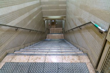 Modern bir metro istasyonunda ya da yeraltı geçidinde temiz bir merdiven manzarası. Bej rengi seramik duvarlar, metal tırabzanlar ve en üstteki dokunsal kaldırımlar.
