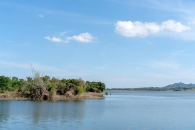 Pofuduk beyaz bulutlu berrak bir gökyüzünün altında sakin mavi bir göl manzarası. Yeşiller yemyeşil ağaçlar ve bitki örtüsü ufuktaki uzak tepelerle kıyı şeridi oluşturur. Huzurlu doğal arkaplan