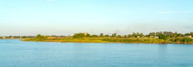 Berrak mavi gökyüzünün altında yemyeşil nehir kıyıları olan sakin bir nehrin geniş panoramik manzarası. Kırsal bir kıyı şeridinde, öğleden sonra ağaçlar ve uzak binalarla küçük bir tekne seyrediyor.