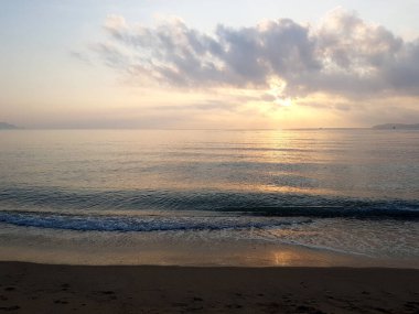 Sakin bir okyanus sahilinde, bulutların arasından süzülen yumuşak altın ışıkla huzurlu bir gün doğumu. Yumuşak dalgalar sabah güneşini yansıtan ıslak kumlara vurur. Huzurlu kıyı manzarası Şafakta hiç insan olmadan
