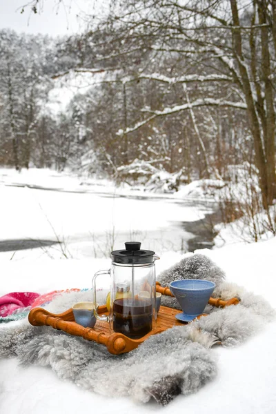 Drinking coffee or tea on the snow in winter with cups, warm blankets and winter landscape with river and forest on background, vertical