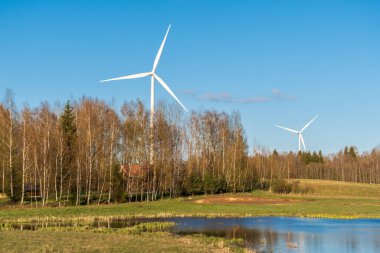 Arka planda açık mavi gökyüzü olan bir orman ve göl yakınında rüzgar türbinleri. Yenilenebilir enerji konsepti, yeşil ekolojik enerji üretimi. Enerji endüstrisi