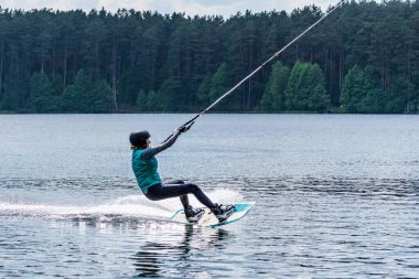 Vilnius, Litvanya - 8 Haziran 2021: Miğfer ve can yeleği olan bir kız arka planda orman, su kaykayı, yazın su sporu ile gölde wakeboard sürüyor 