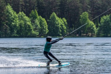 Vilnius, Litvanya - 8 Haziran 2021: Miğfer ve can yeleği olan bir kız arka planda orman, su kaykayı, yazın su sporu ile gölde wakeboard sürüyor 