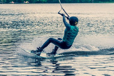 Vilnius, Litvanya - 8 Haziran 2021: Miğfer ve can yeleği olan bir kız arka planda orman, su kaykayı, yazın su sporu ile gölde wakeboard sürüyor 