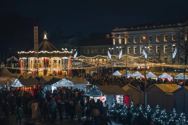 Vilnius, Litvanya - 30 Kasım 2025: Vilnius, Litvanya, Avrupa 'da, insanlar, tatil ışıkları ve pazar tezgahlarıyla çevrili bir şehir meydanında gece güzel bir atlıkarınca