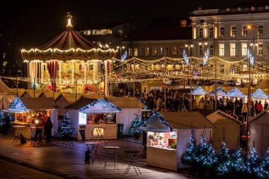 Vilnius, Litvanya - 3 Aralık 2025: Vilnius, Litvanya, Avrupa 'da, insanlar, tatil ışıkları ve pazar tezgahlarıyla çevrili bir şehir meydanında gece güzel bir atlıkarınca