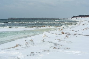 Kış mevsiminde karla kaplı kum tepeleri sahilde yürüyen birkaç kişi ve Litvanya, Palanga 'da bir rıhtım.