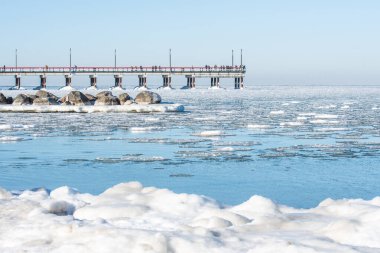 Durgun kış denizi, solgun bir gökyüzünün altında yüzen buz kütleleriyle ve arka planda Palanga ahşap köprüsü ya da iskelesiyle benekli.