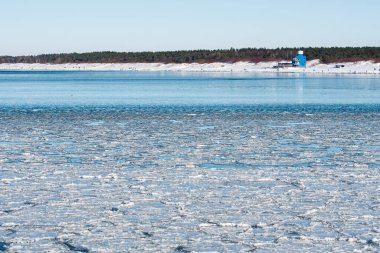 Durgun kış denizi solgun bir gökyüzünün altında yüzen buz kütleleriyle kaplı. Arka planda kar ve ormanla kaplı kum tepeleri var.