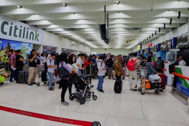 Havaalanı terminali sahnesinde bavullar ve bagaj arabalarıyla havayolu gişelerinde sırada bekleyen bir sürü yolcu var..