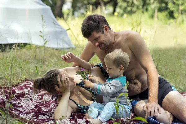 Jonge vader, moeder en zoon 1.3 jaren een picknick op het gras te liggen. Zoon geeft moeder wilde bloemen. — Stockfoto