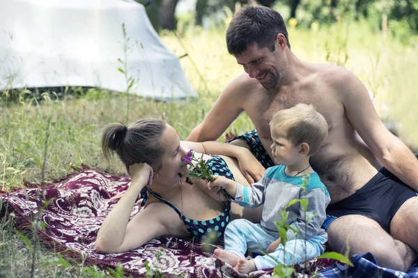 Jonge vader, moeder en zoon 1.3 jaren liggen op het gras in picknick. Zoon geeft moeder wilde bloemen. — Stockfoto