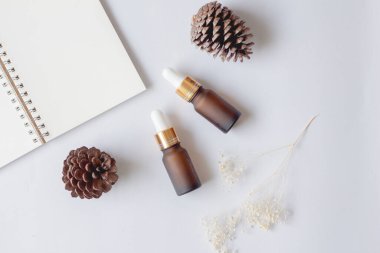 Düz organik aromaterapi yağları ya da cilt bakım ürünleri. Not defterli damlatan cam şişe, kozalak ve beyaz arka planda kuru çiçek..