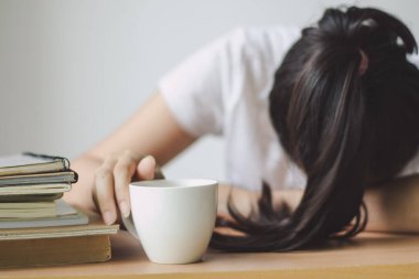 Elinde bir fincan kahve tutan yorgun genç bir kadın kitap ve iş ile masanın üzerinde yatıyor..