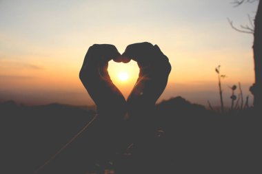 Gün batımında dağda güneş batarken kalp şeklinde el ele tutuşan kadın..
