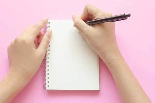 女人的手在粉红桌上的空笔记本上写字 平面放置顶部视图 — 图库照片