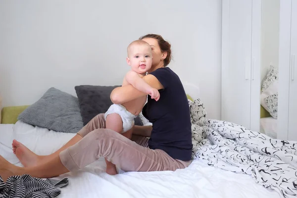 Mom dresses the baby on the bed in the bedroom in striped pajamas. Toddler looks into the frame